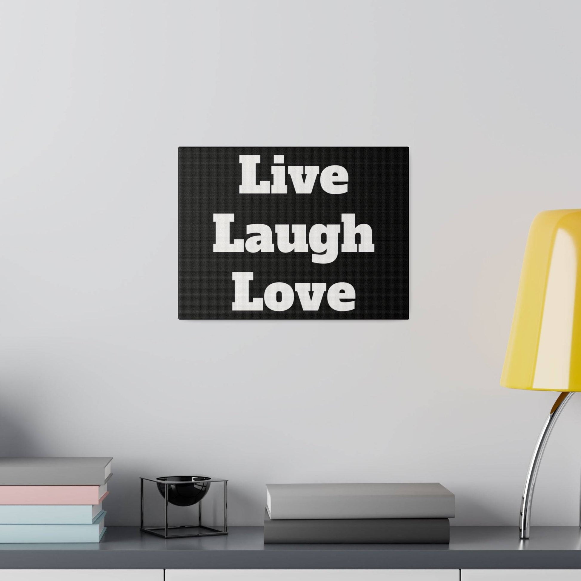 Black wall art with white Live Laugh Love text above a shelf holding pastel books, a small black metal bowl, and a yellow lamp.