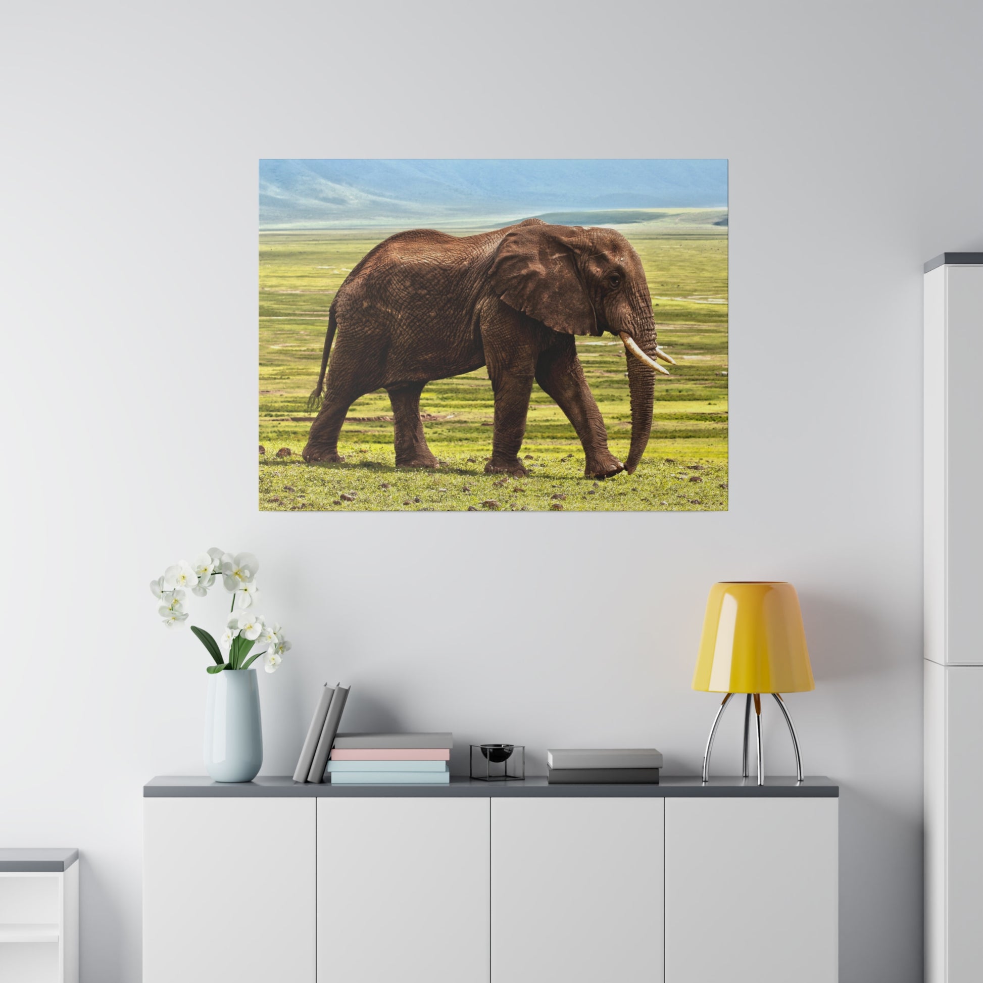 Brown elephant walking across a grassy savannah in a framed print mounted on a light gray wall above a white cabinet with a yellow lamp.
