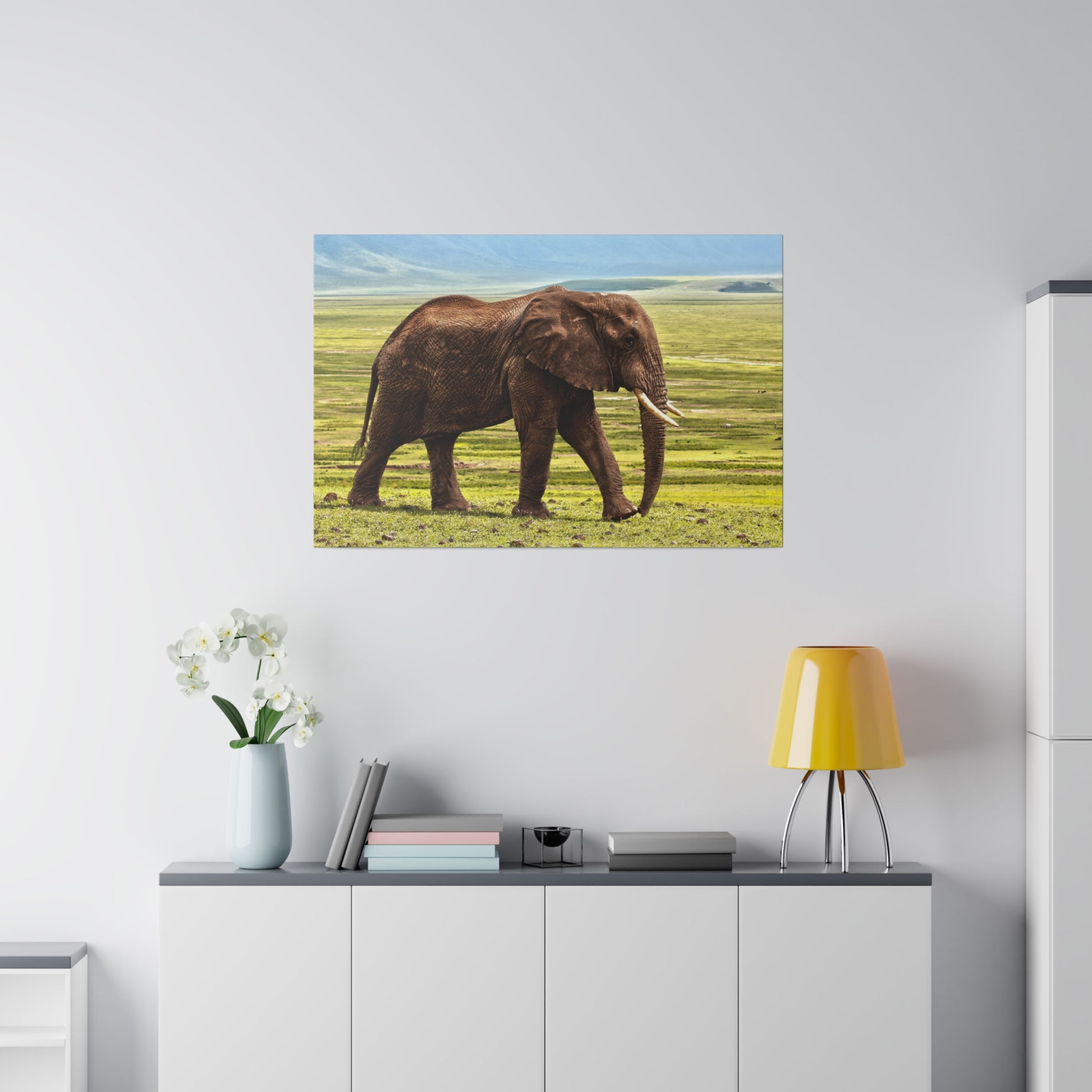 Brown African elephant walking on grassy plains with blue sky in a wall-mounted photo above a white cabinet.