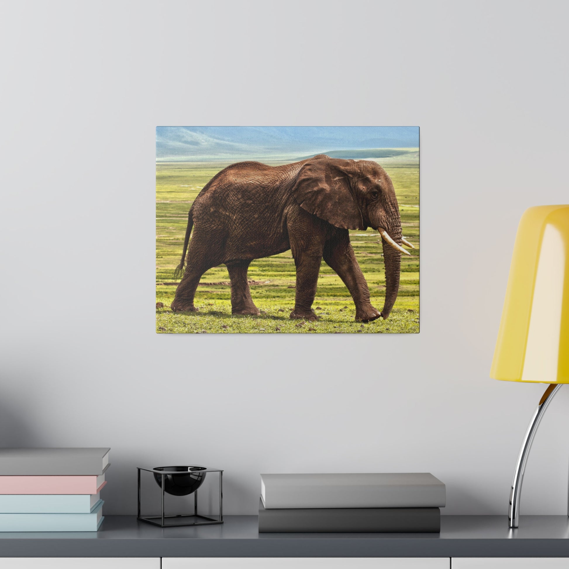 Brown African elephant walking on a grassy savanna beneath a blue sky, depicted on a square canvas mounted on a light gray wall.
