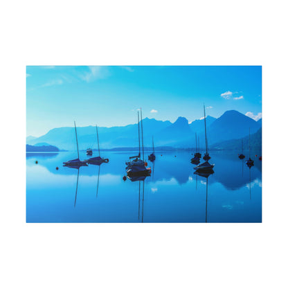 Calm blue lake with several anchored sailboats, their masts mirrored in the water, mountains in the distant background.