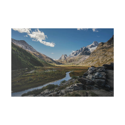A winding river runs through a green valley flanked by jagged snow‑capped mountains under a blue sky.