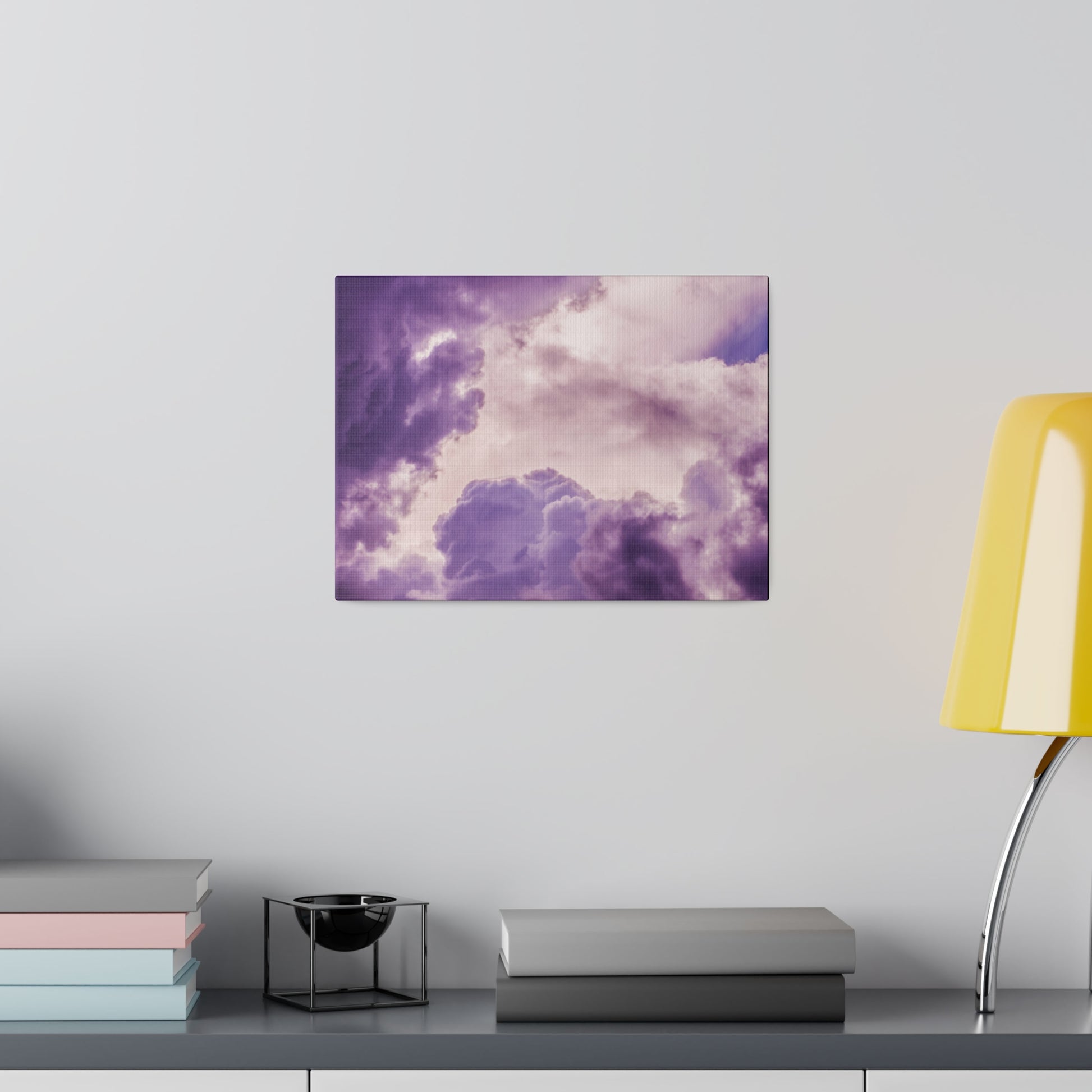 Purple cloud-themed wall art hangs above a shelf with stacked books, a black metal candle holder, and a yellow lamp.