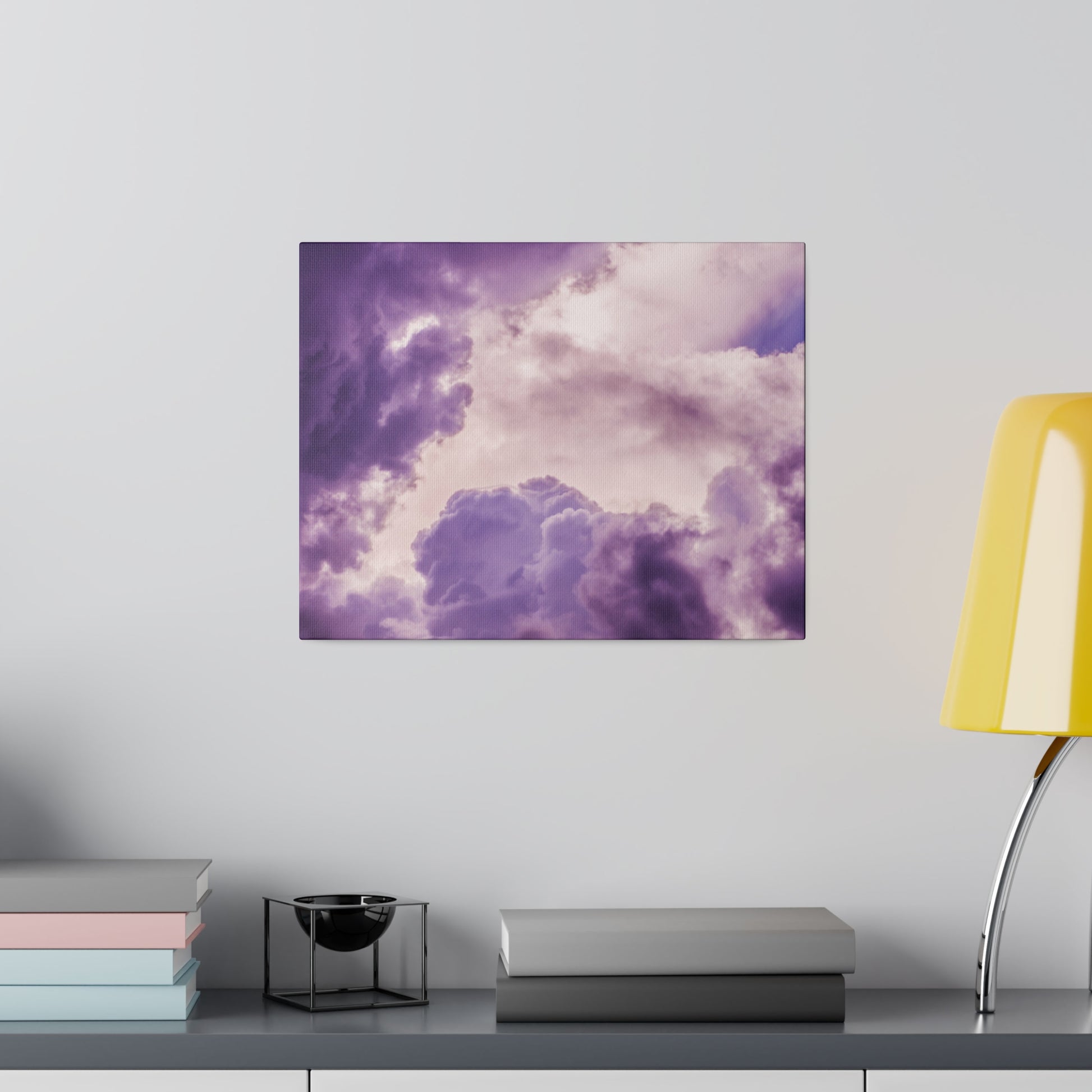Purple cloud-themed canvas print on a gray wall above a shelf with stacked books, a small black bowl, and a yellow lamp.