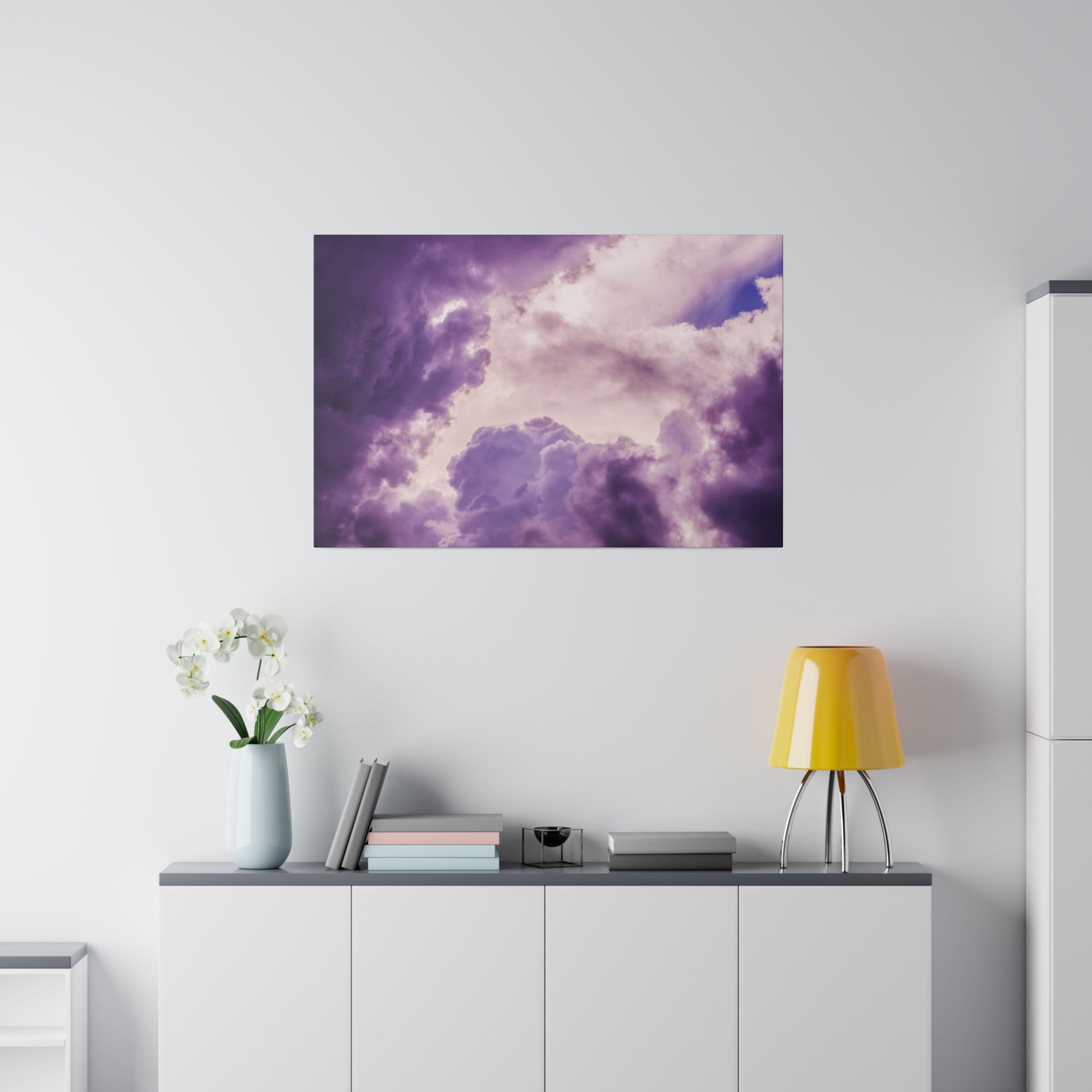 Purple cloudscape art print hanging above a white sideboard with a light blue vase of white orchids and a yellow lamp.