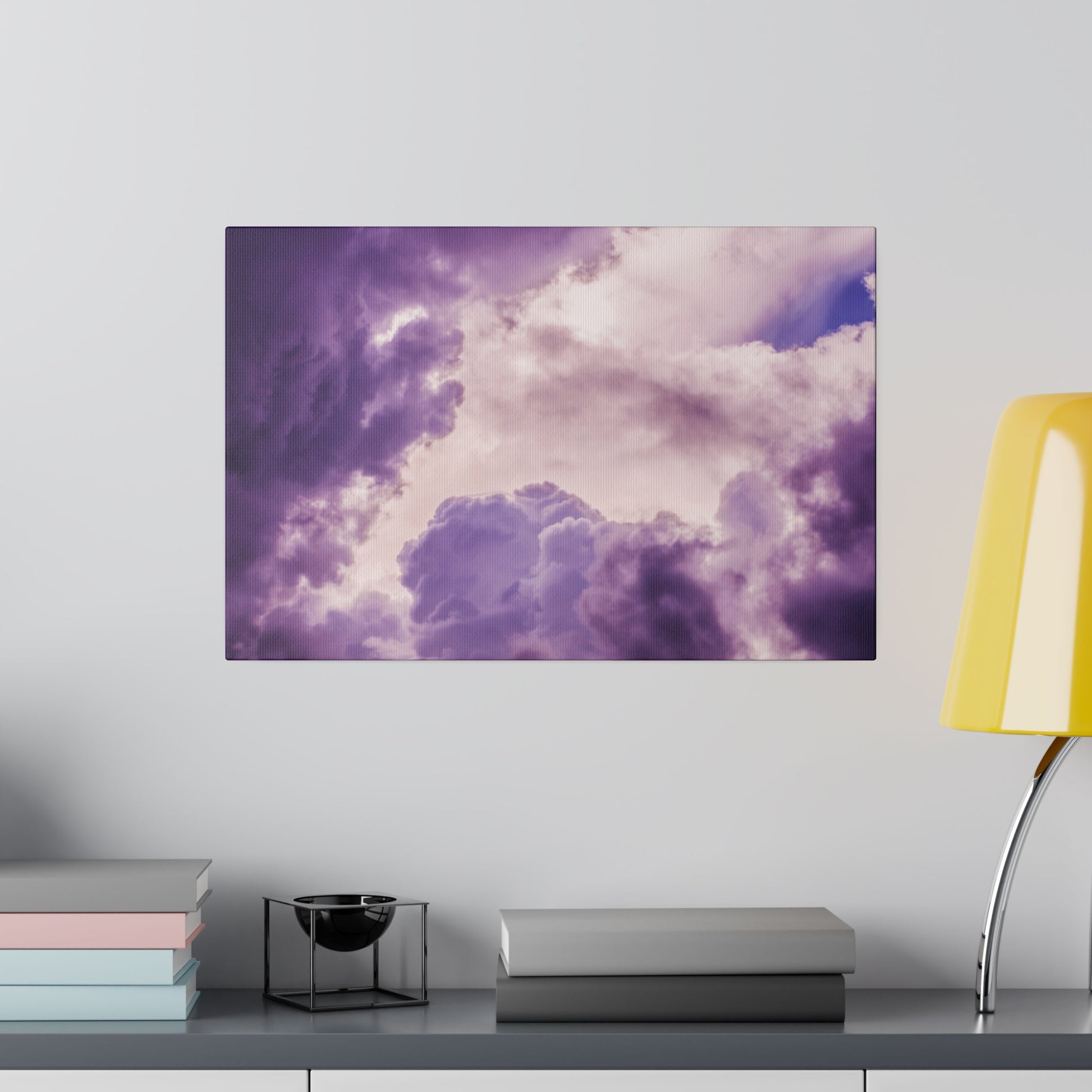 Rectangular wall art depicting purple and pink clouds hangs above a desk with stacked pastel books and a yellow lamp.