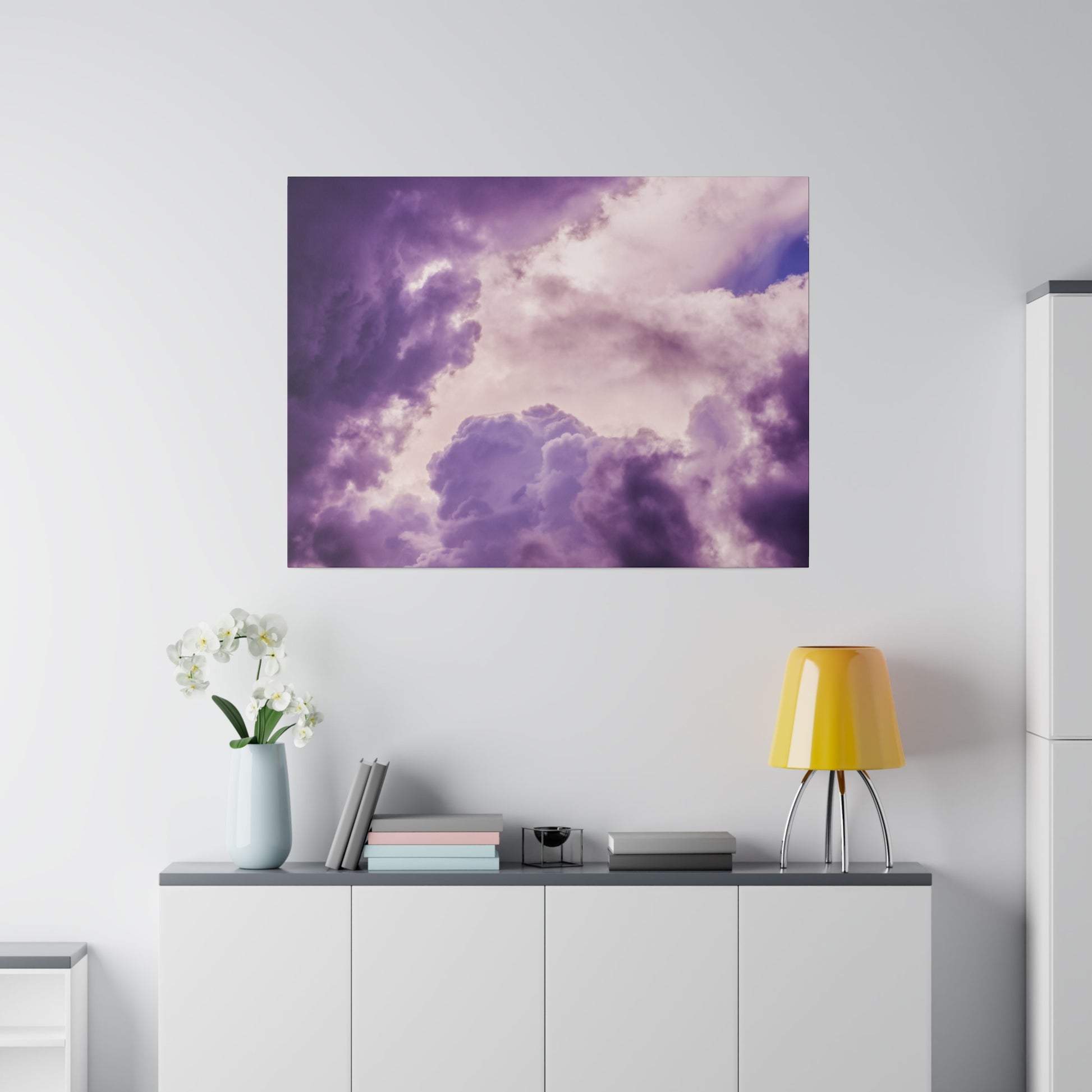 Square wall art of purple and pink clouds mounted above a white sideboard with a pale blue vase, books, and a yellow table lamp.