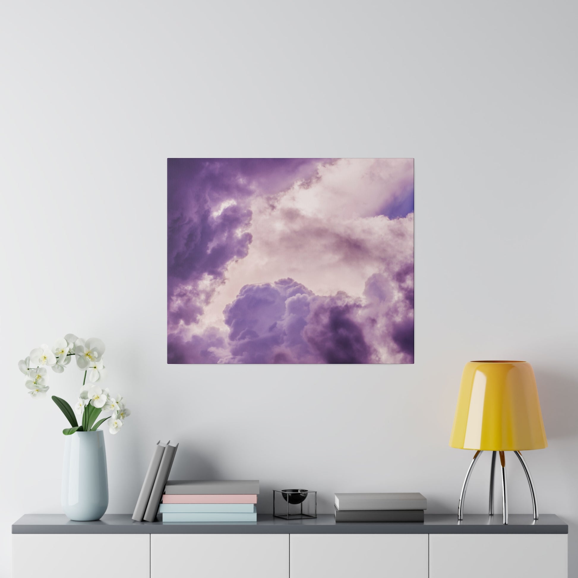 Purple-toned cloud artwork mounted on a light wall above a console with a pale blue vase of white orchids, stacked books, and a yellow