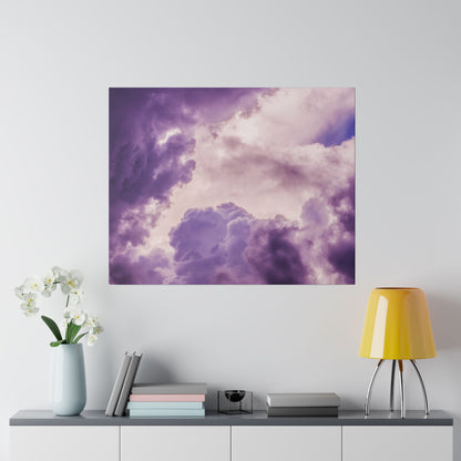 Square wall art print depicting purple and pink cloudy sky hung above a gray console table with a light blue vase, books, and a yellow lamp.