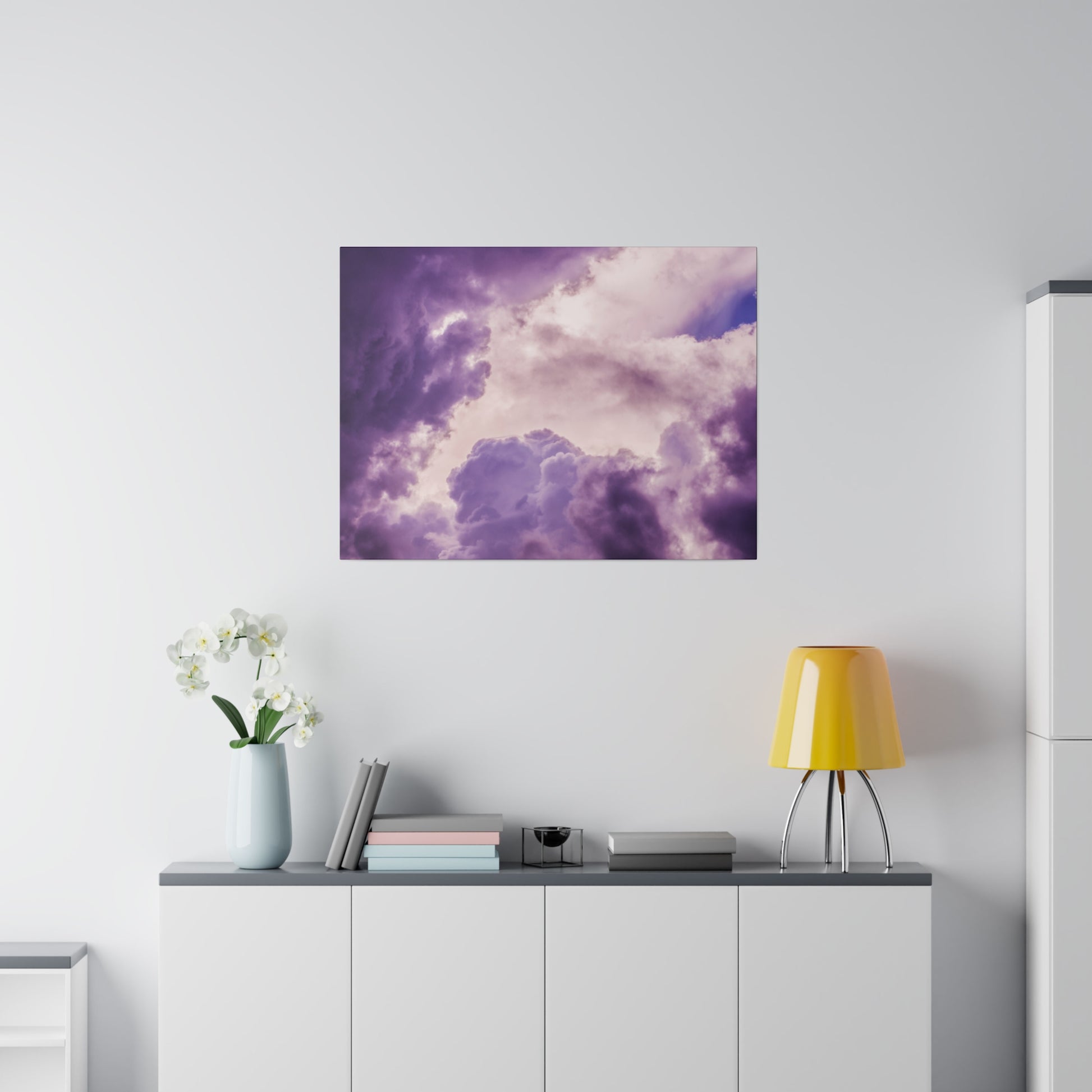 Square wall art depicting purple cloudy sky hangs above a white sideboard with a yellow lamp and a vase of white orchids.