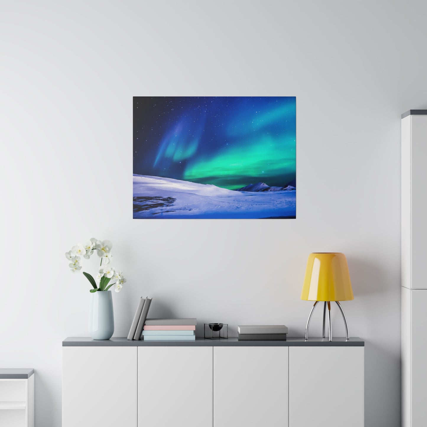 Square wall art of green and blue aurora over a snowy landscape, hung above a white cabinet in a modern room.