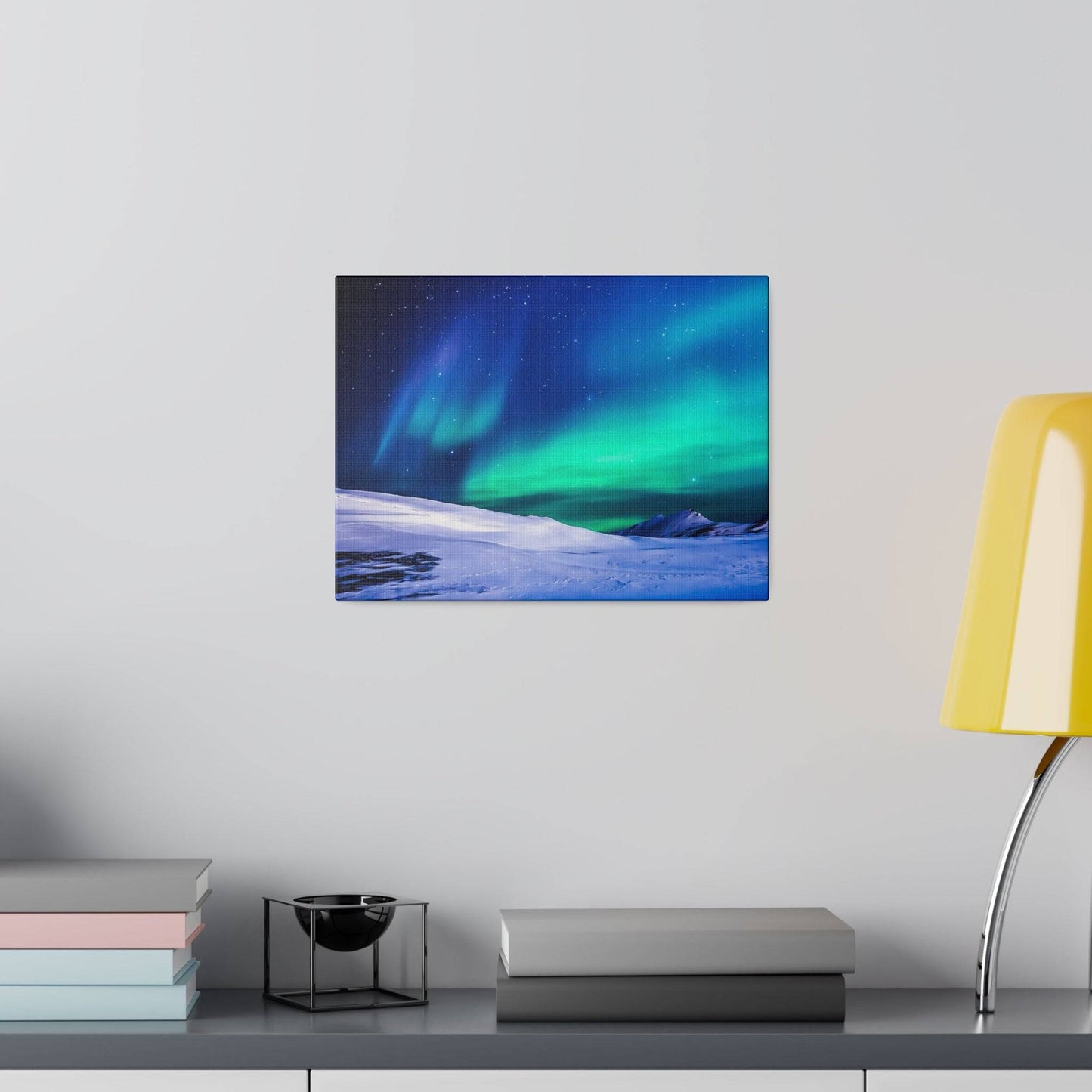 Mounted canvas print of a snowy landscape with green and blue Northern Lights on a gray wall above a desk with books and a yellow lamp.
