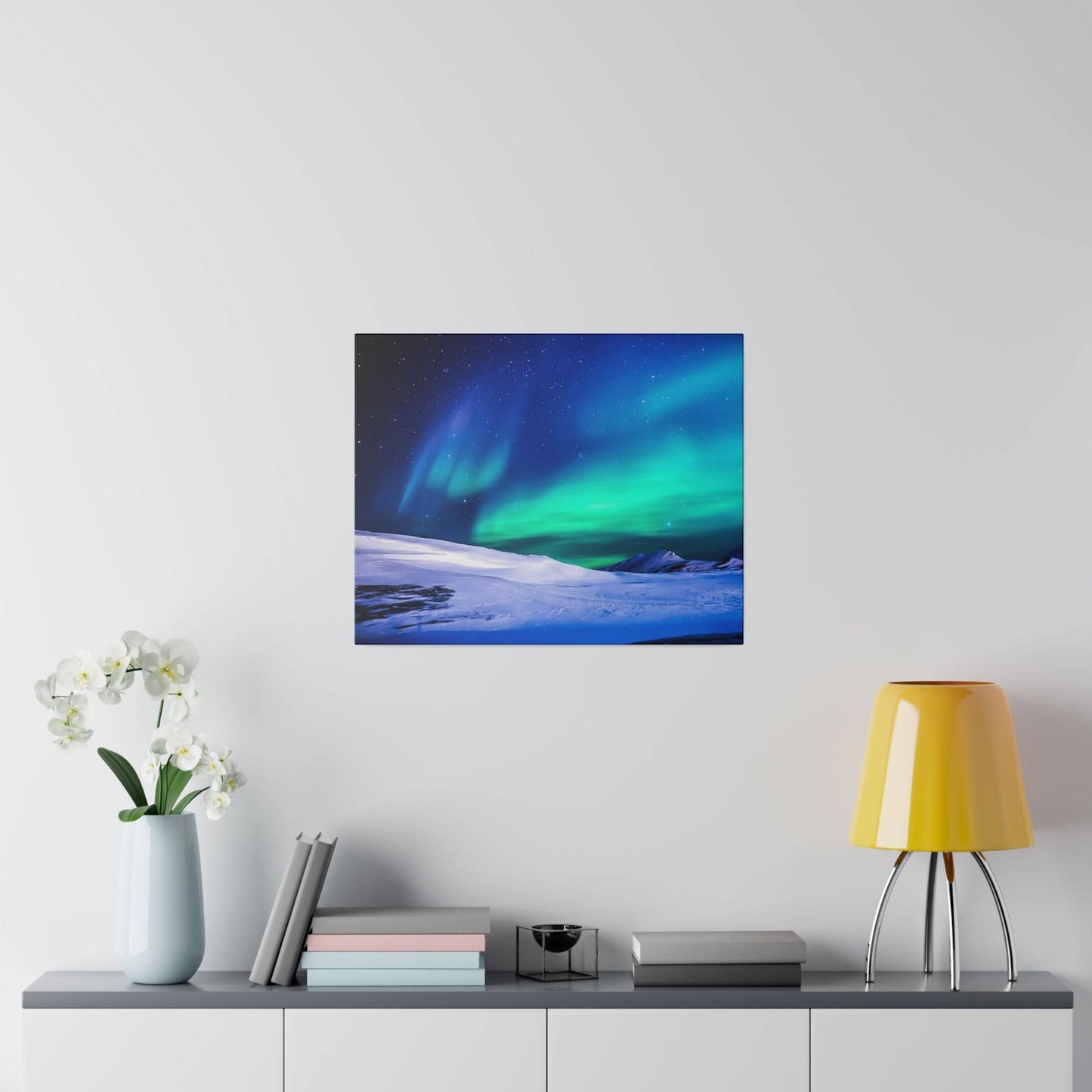 Square print of a snowy landscape with green and blue aurora borealis in a dark night sky, hung on a light gray wall above a console.