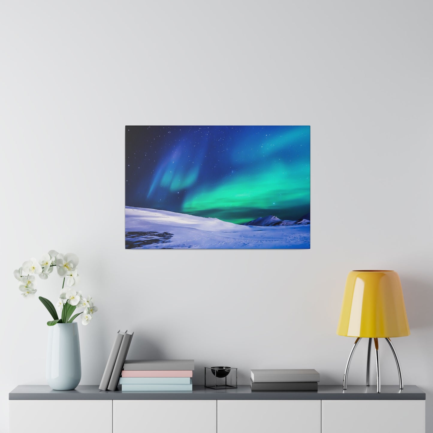 Framed landscape photo of the northern lights over snow hangs on a white wall above a sideboard with a vase of white orchids, books, and a