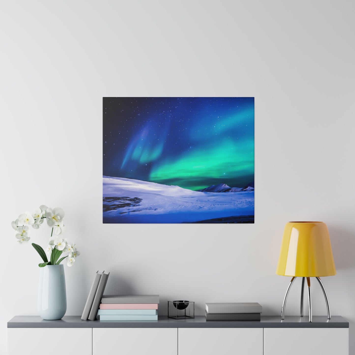 Square canvas of the aurora borealis over snowy hills in blue and green hues, mounted on a light gray wall.