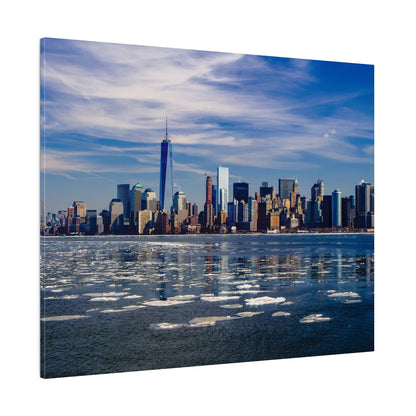 New York City skyline across an icy river, with One World Trade Center amid tall buildings under a blue sky.