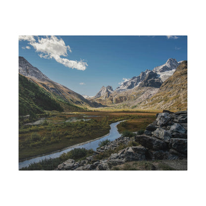Alpine valley with a winding river through a grassy meadow, surrounded by snow-capped mountains and a rocky foreground.