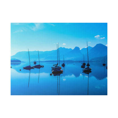 Calm blue lake with several small sailboats anchored, reflections on the water and a distant mountain range.