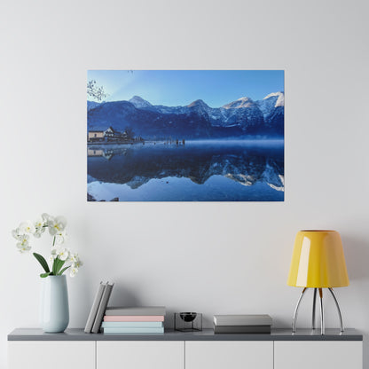 Photograph of a snow-capped mountain lake with a small building and dock reflected in calm blue water.