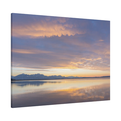 Calm lake at sunset with orange and purple sky reflecting on gentle water, distant snow-capped mountains.