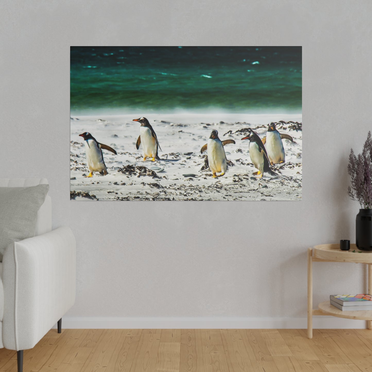 A group of penguins near the ocean, walking on a sandy beach. The green-blue sea provides a serene backdrop, with scattered seaweed adding to the natural setting.