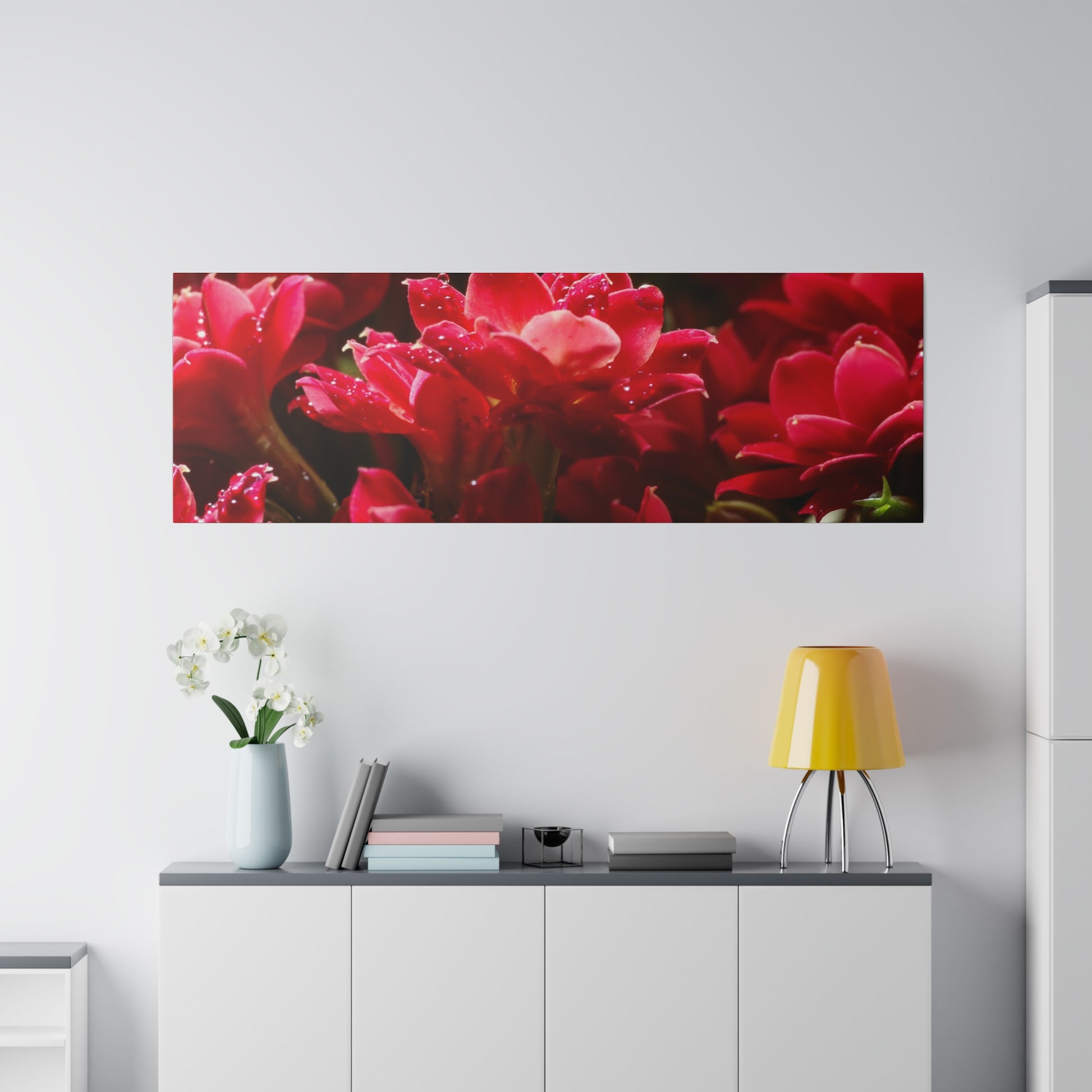 Rectangular wall art of red flowers hangs above a white cabinet, with a pale vase of white orchids and a yellow table lamp.