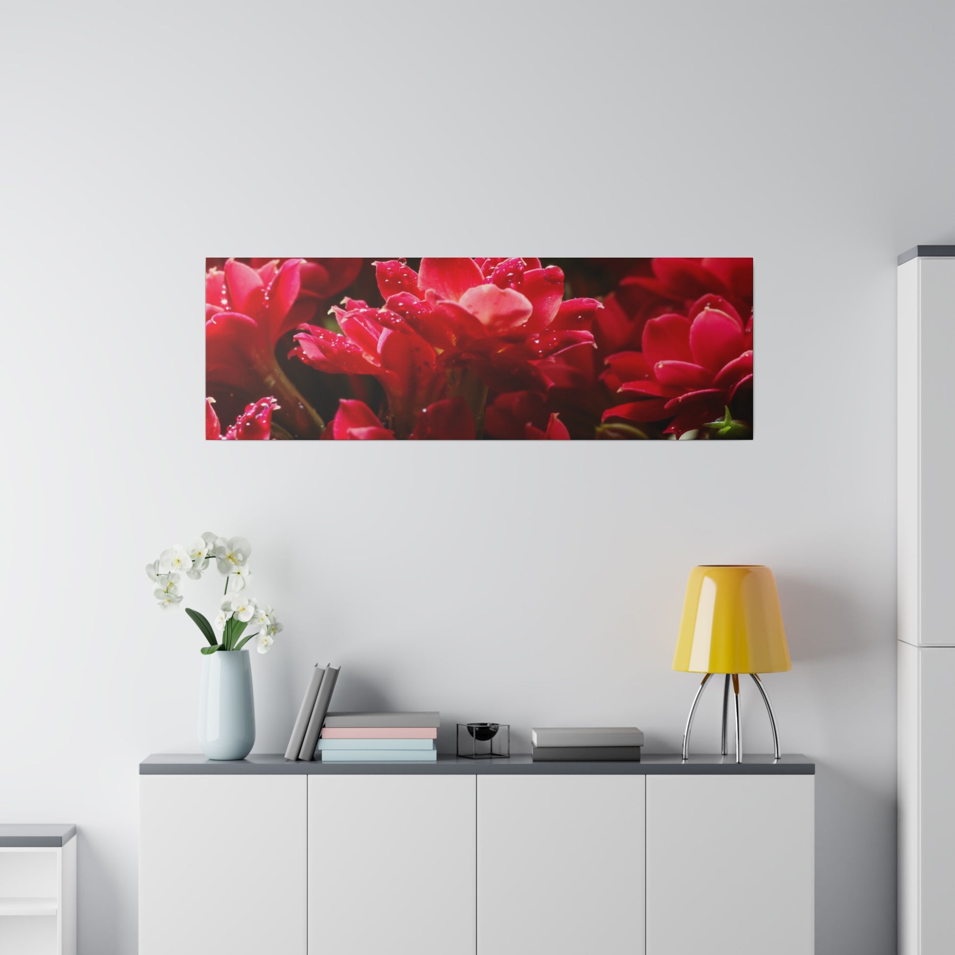 Rectangular wall canvas showing a close-up of red flowers above a white cabinet with a yellow lamp and vase.