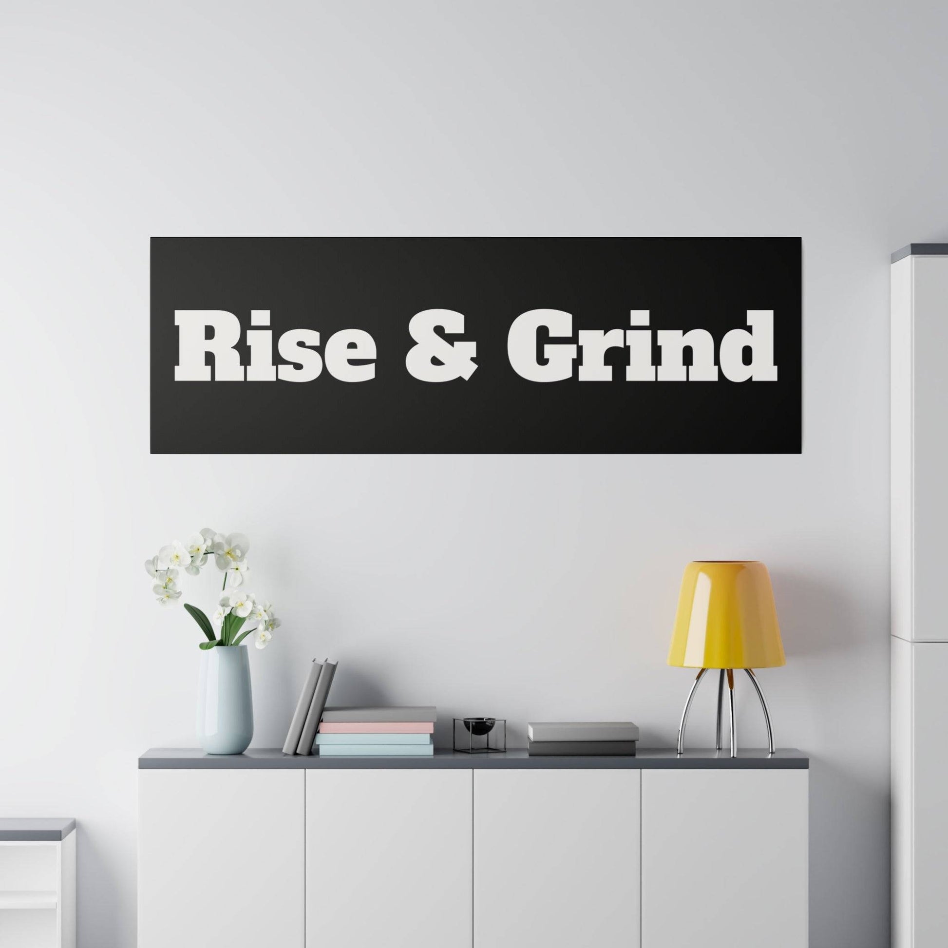 Black rectangular wall sign reading "Rise & Grind" in bold white letters above a white console with a yellow lamp.