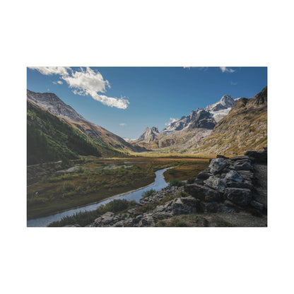 Snow-dusted mountains rise over a green valley with a winding river and a rocky foreground.