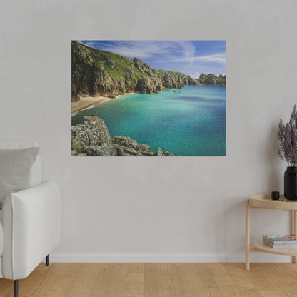 Square photo of a rocky coastal cove with turquoise sea, sandy beach, and green cliffs under a blue sky.