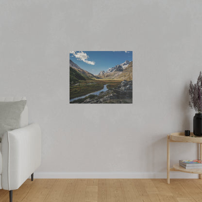 Square landscape photo hung on a light gray wall depicting a mountain valley with a winding river and snow-capped peaks.