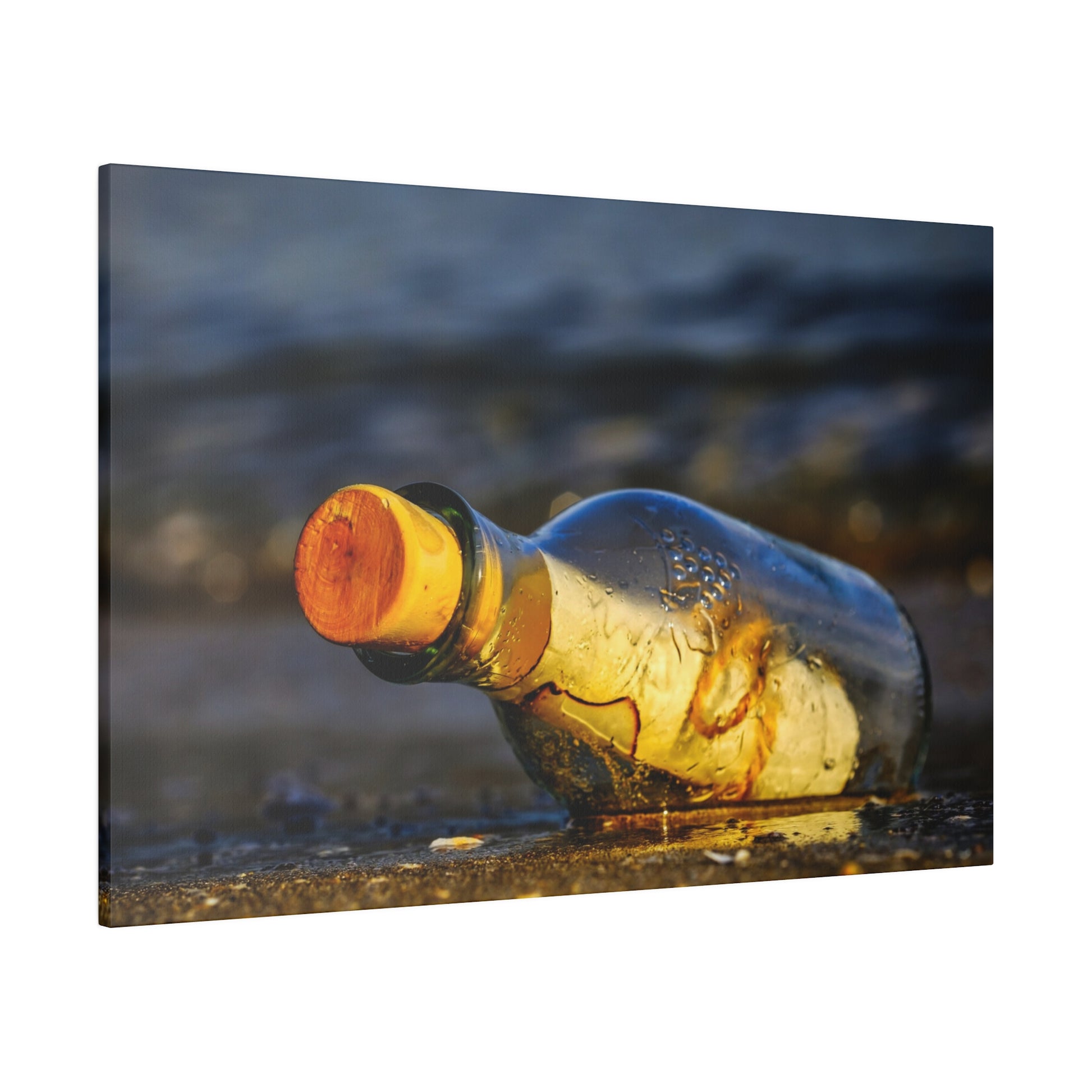 A clear glass bottle with an orange cork lies on wet sand near the shoreline, with water in the background.