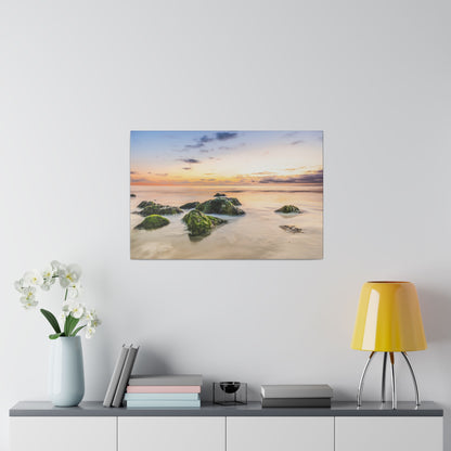 A sunset seascape painting showing orange and purple sky, calm water, and moss-covered rocks in the foreground.