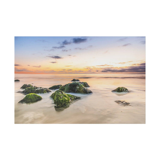 Colorful sunset over a calm sea with moss-covered rocks protruding from the shallow water.