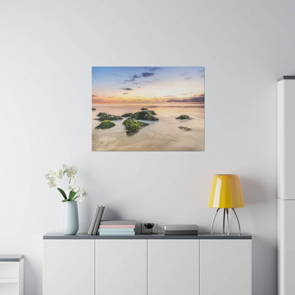 A seascape painting of a sunset over a calm beach with mossy rocks in the foreground.