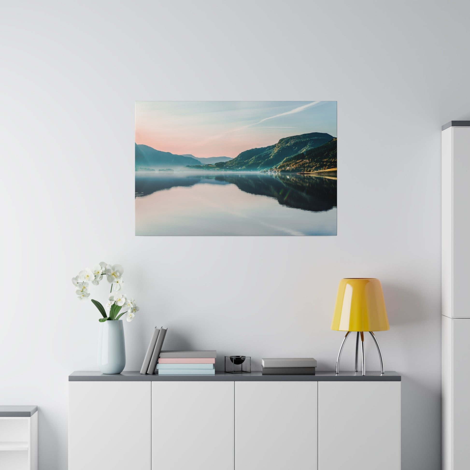 Landscape photograph of a calm lake and mountains at sunset, hanging on a white wall above a cabinet with a blue vase, books, and a yellow
