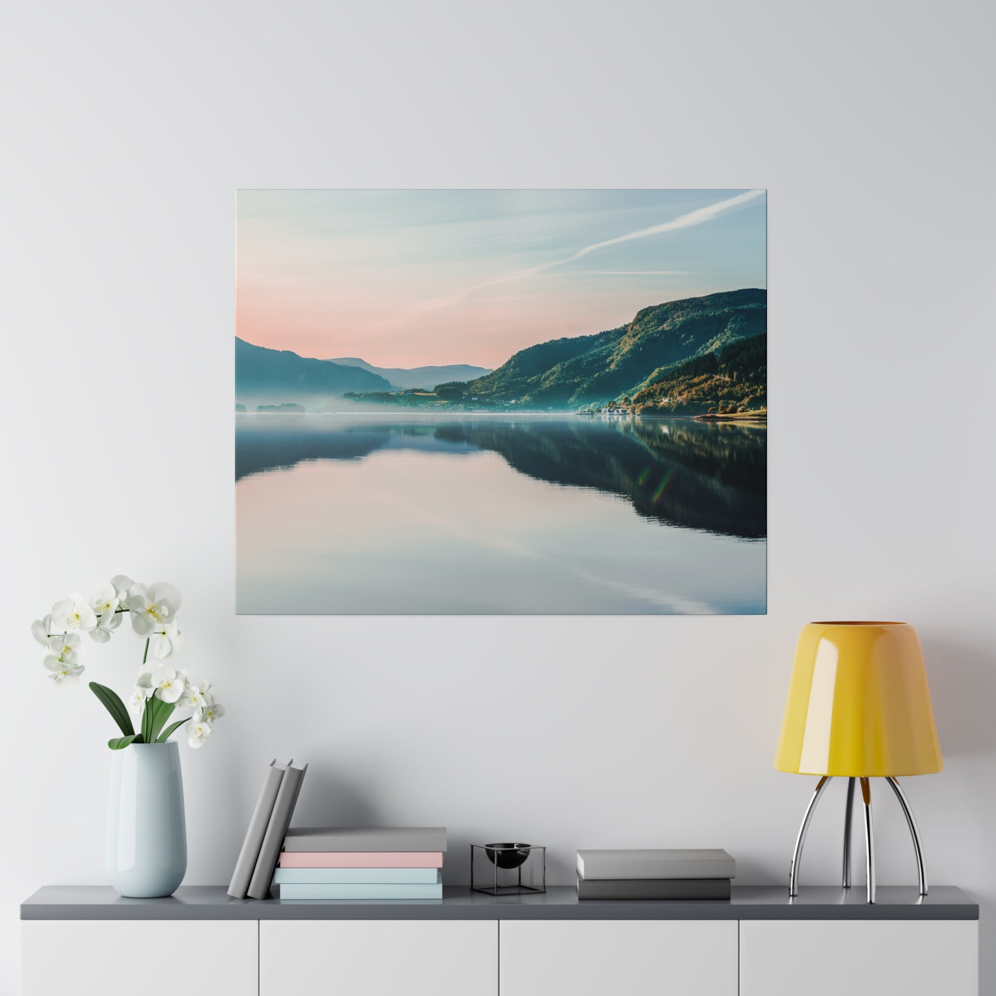 Landscape photo of a calm lake, mountains, and pastel sky reflected in the water, mounted above a sideboard with a vase and a yellow lamp.