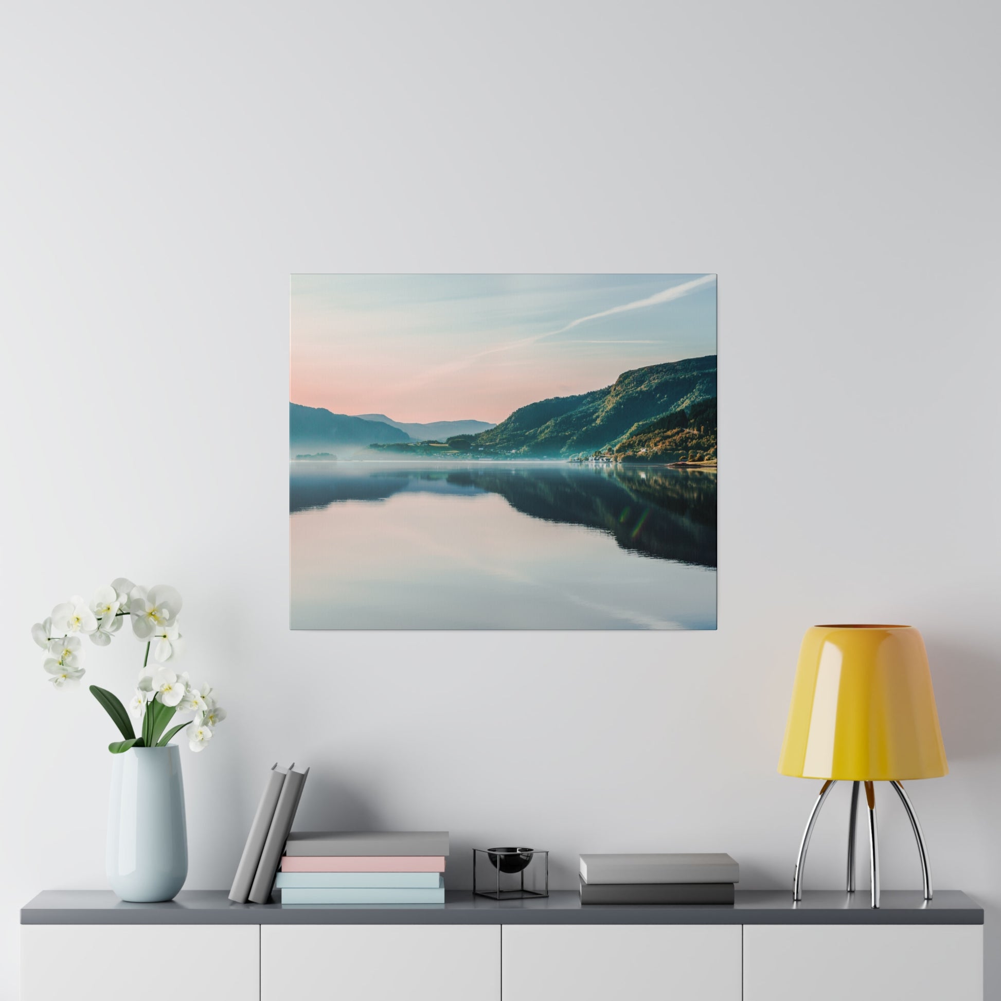 Square wall art of a calm lake, mountains, and pastel sky with reflection; hung above a gray cabinet with white orchids and a yellow lamp.