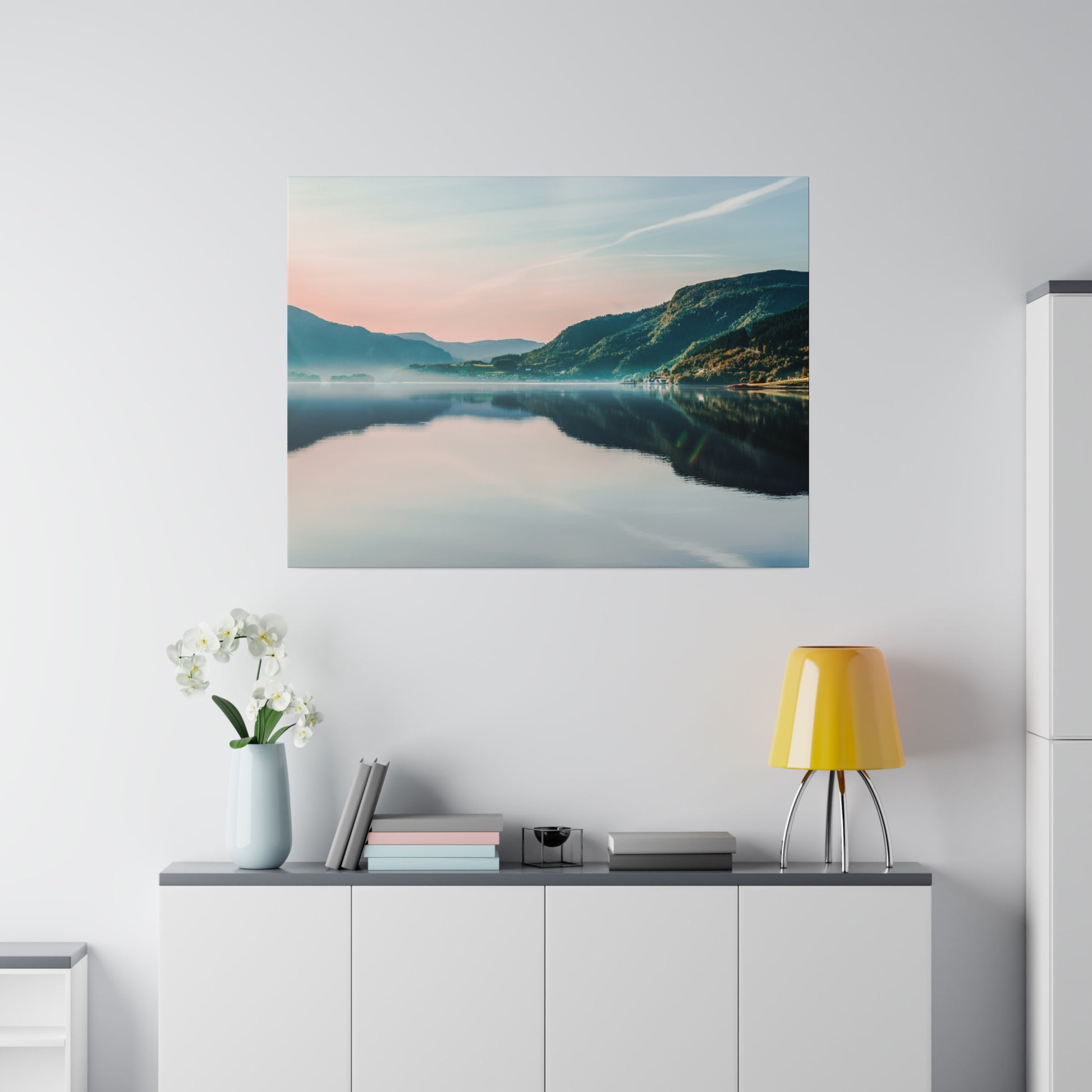 Square wall art of a serene lake and mountains at sunset, mounted above a white cabinet with a yellow lamp and white orchid vase.
