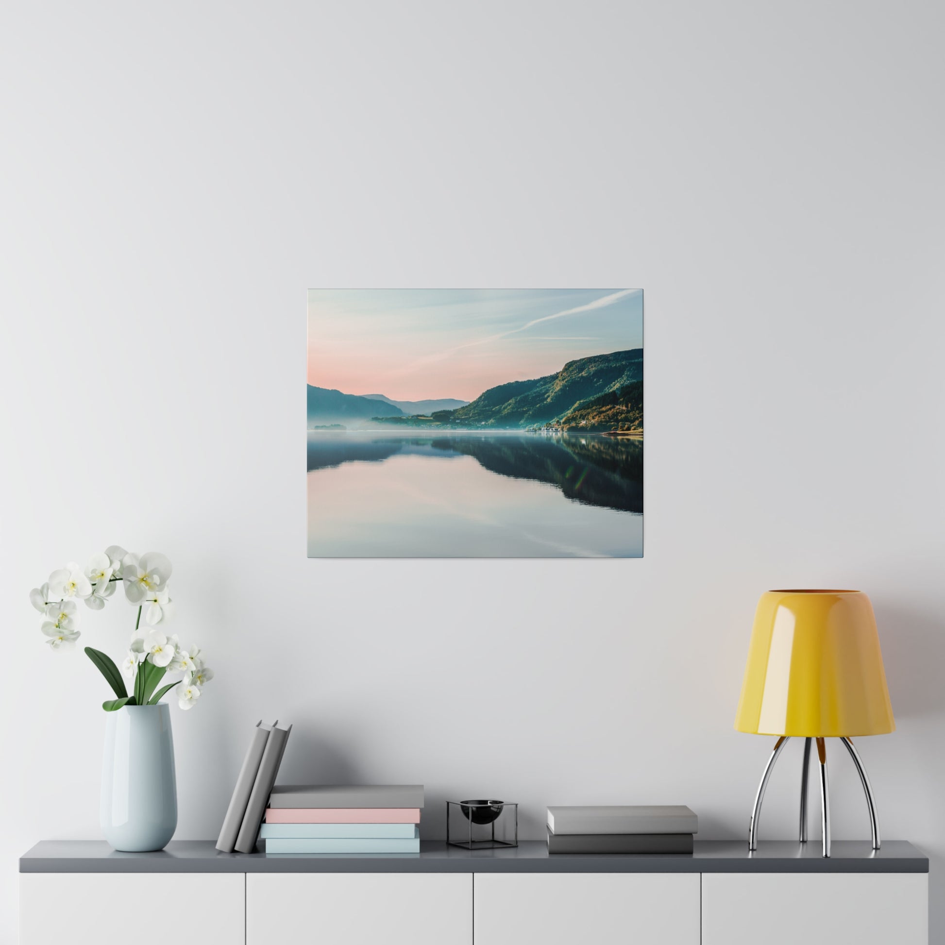 Framed landscape photograph of a calm lake and mountains at sunrise, hung above a gray sideboard.