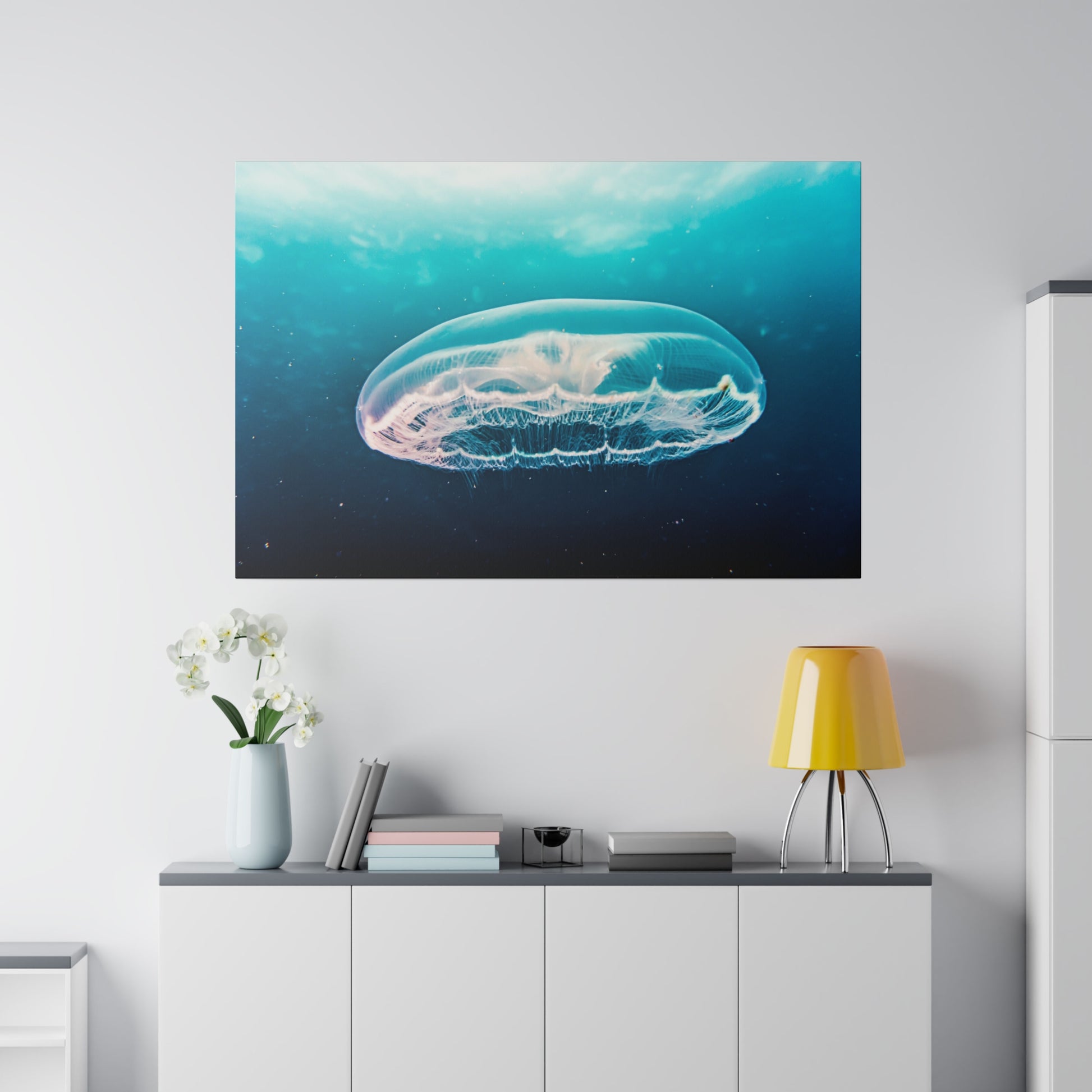 Framed wall art of a translucent jellyfish glowing white and pink in a blue ocean, hanging above a white cabinet.