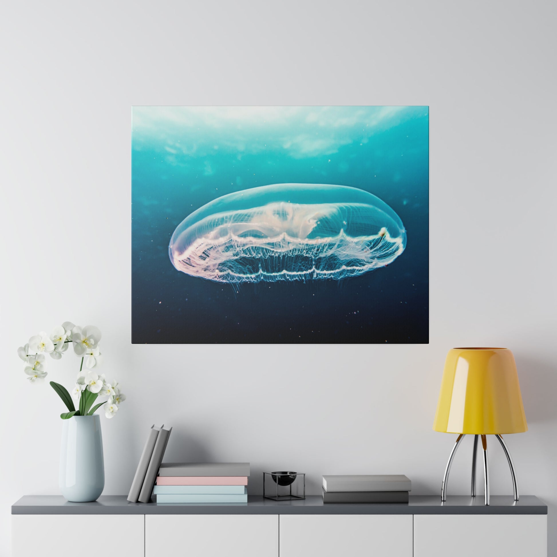 Square wall art of a translucent jellyfish floating in blue-green ocean, mounted above a console table with a vase and yellow lamp.