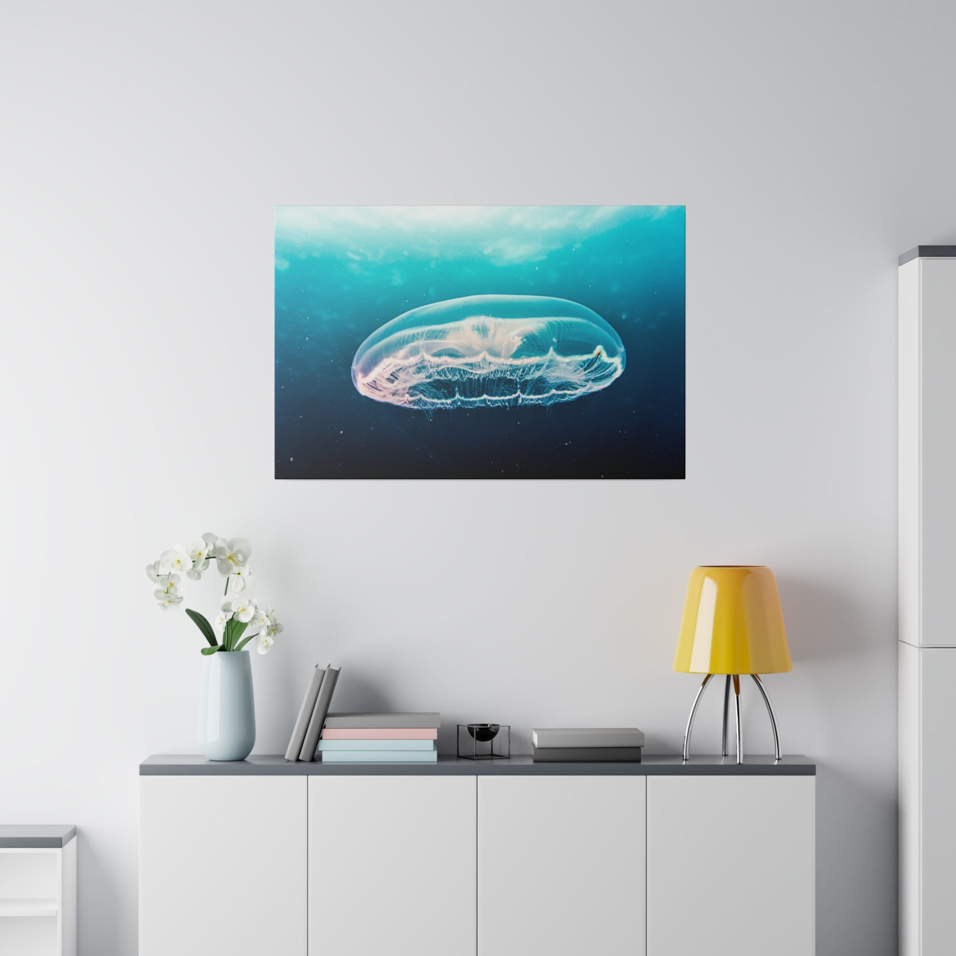 Underwater jellyfish canvas hangs above a white sideboard with a yellow lamp, books, and a vase of white flowers.