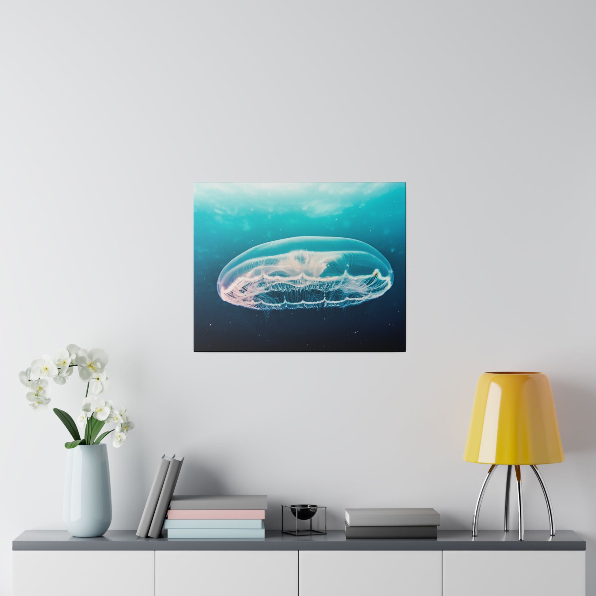 Square wall art depicting a transparent jellyfish drifting underwater, mounted above a gray sideboard with books and a yellow lamp.