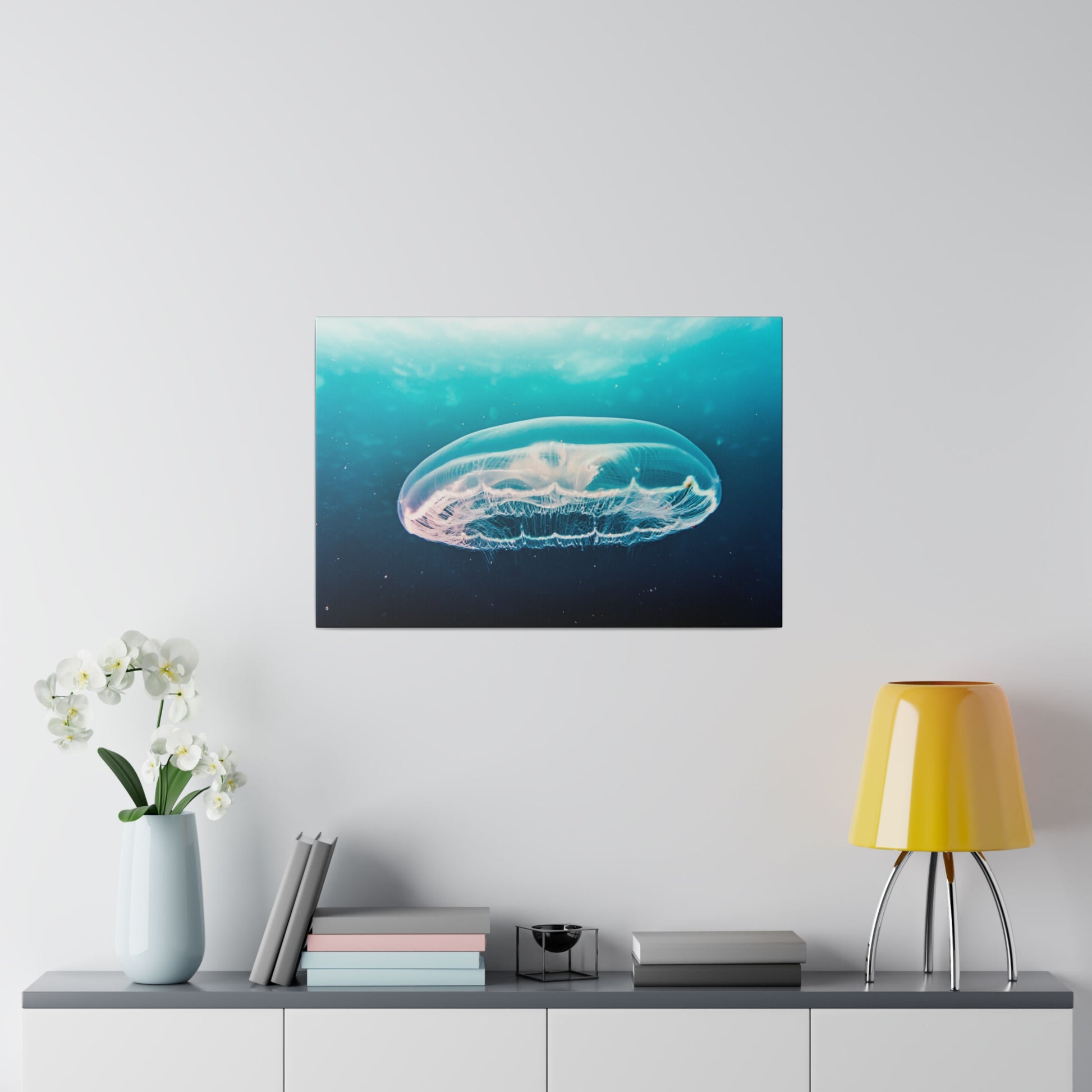 Framed underwater photograph of a translucent jellyfish in turquoise water, hung on a light wall above a cabinet with a vase, books, and a