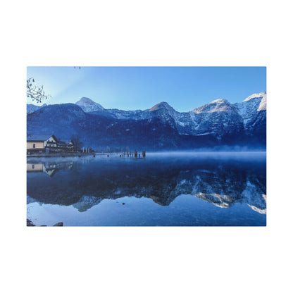 Snow-capped mountains rise beyond a calm blue lake with a small dock and buildings reflected on the water.