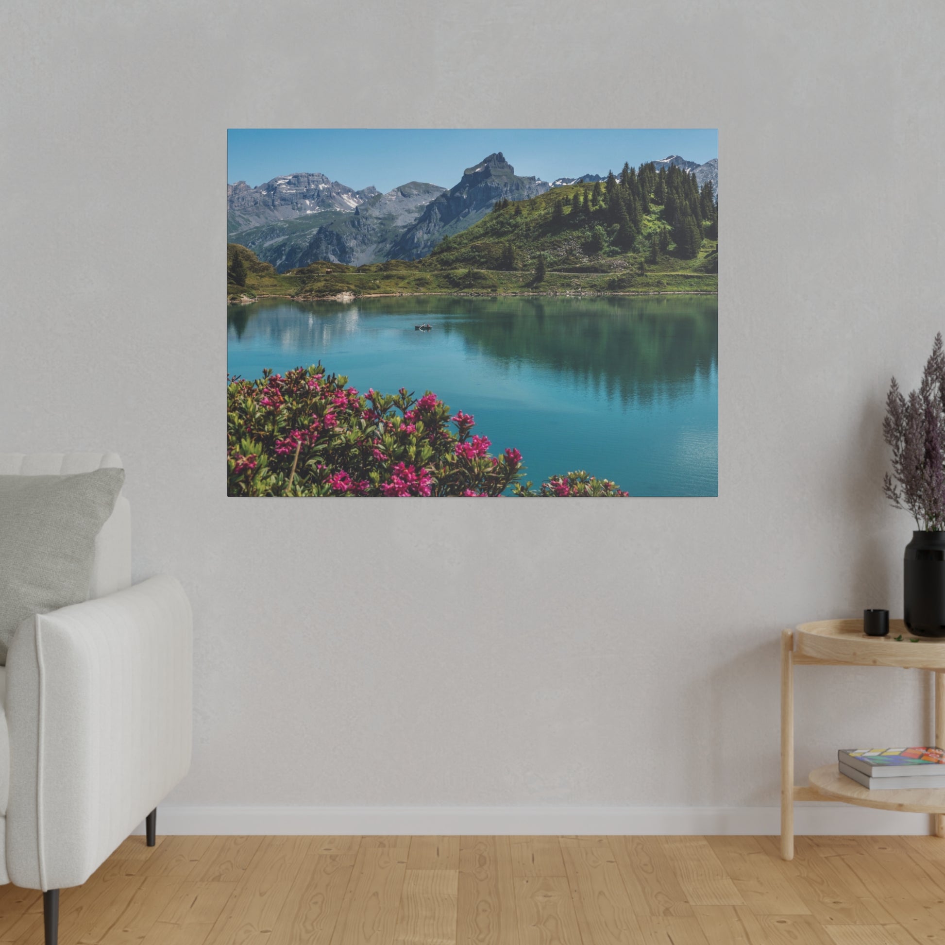 Landscape photograph of a turquoise lake surrounded by green hills and snow-capped mountains, with pink flowers in the foreground.