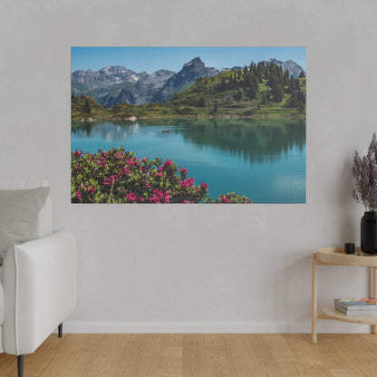 Landscape photograph of a turquoise lake surrounded by green hills and rugged mountains, with pink flowers in the foreground.