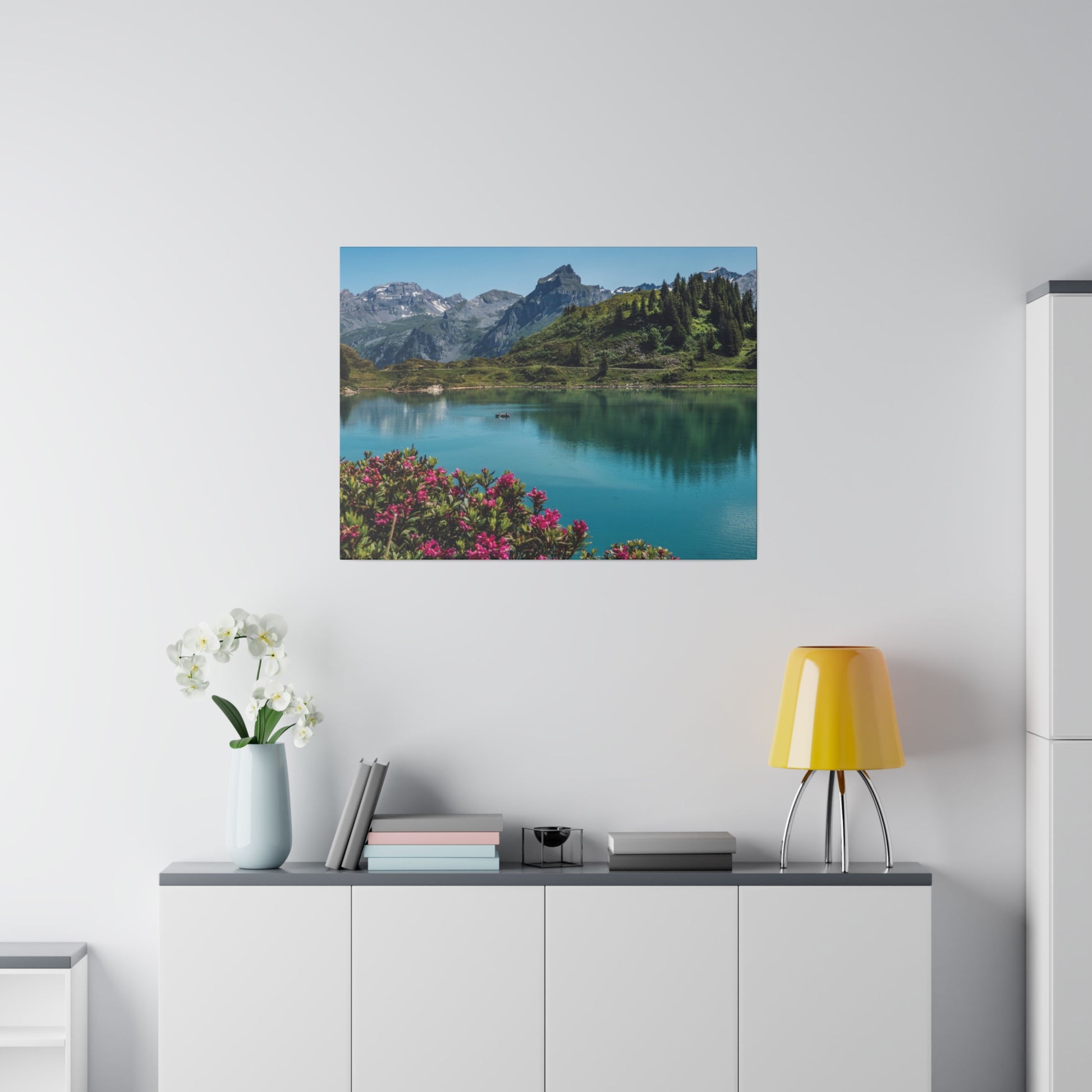 Square wall art depicting a turquoise lake, mountains, and pink foreground flowers above a white cabinet in a minimal room.