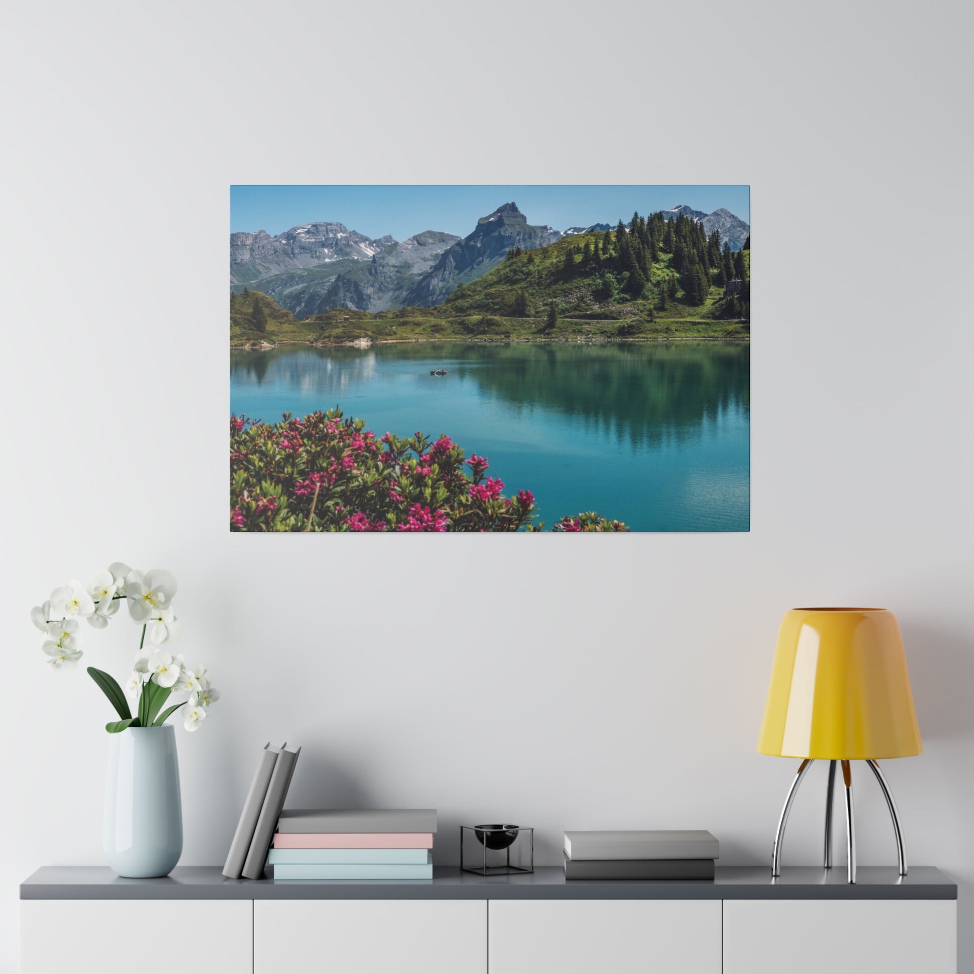 Landscape photograph of a turquoise lake, green hills and mountains with pink flowers in the foreground, mounted on a white wall.